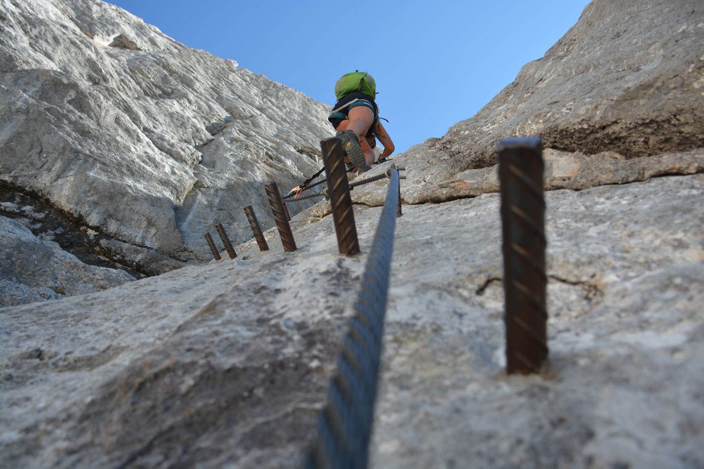 Eisenstifte ragen in einer Reihe aufwärts aus der Felswand, weiter oben klettert eine Person am Seil. 