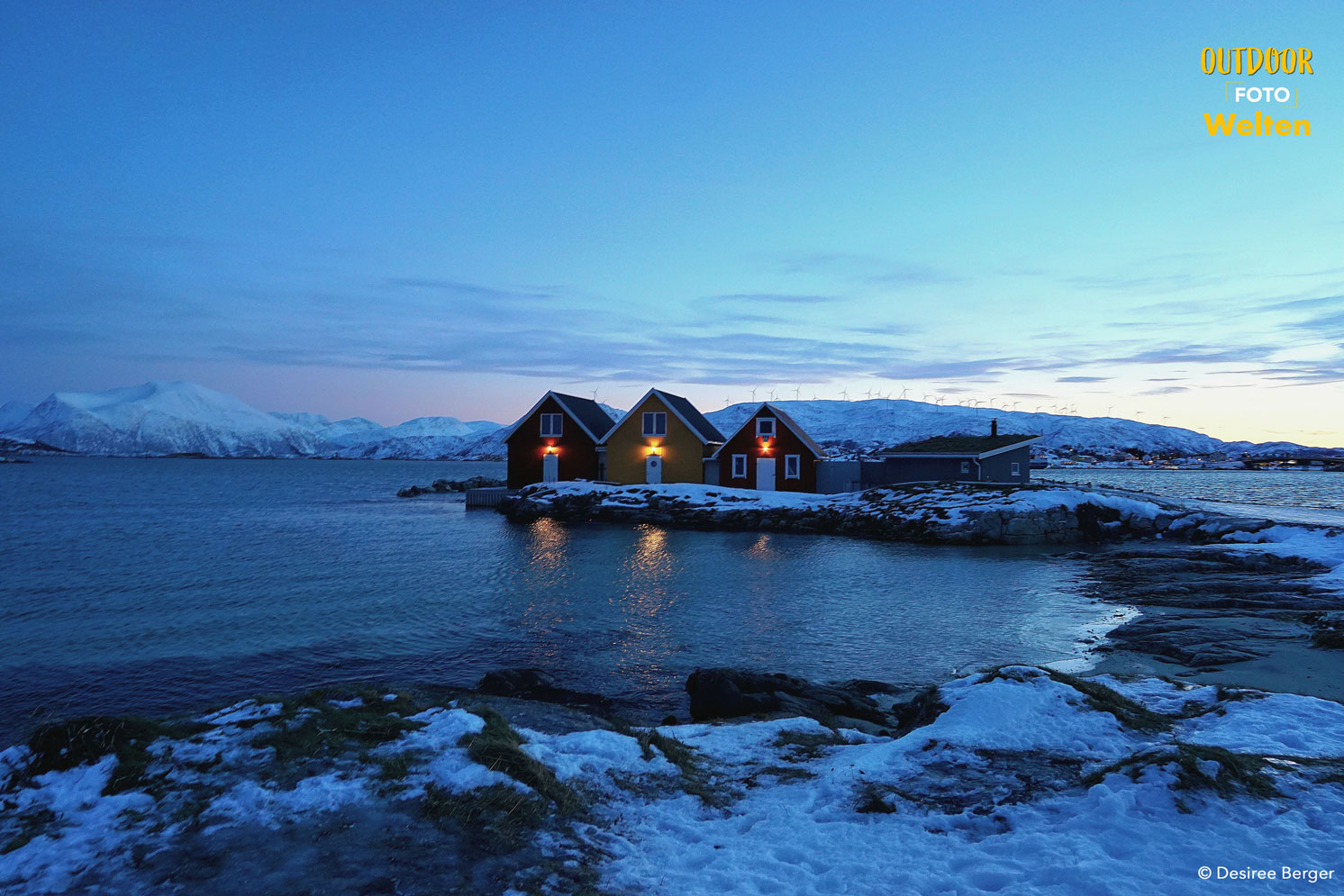 Drei kleine Häuser stehen am Wasser, dahinter schneebedeckte Hügel und der Abendhimmel