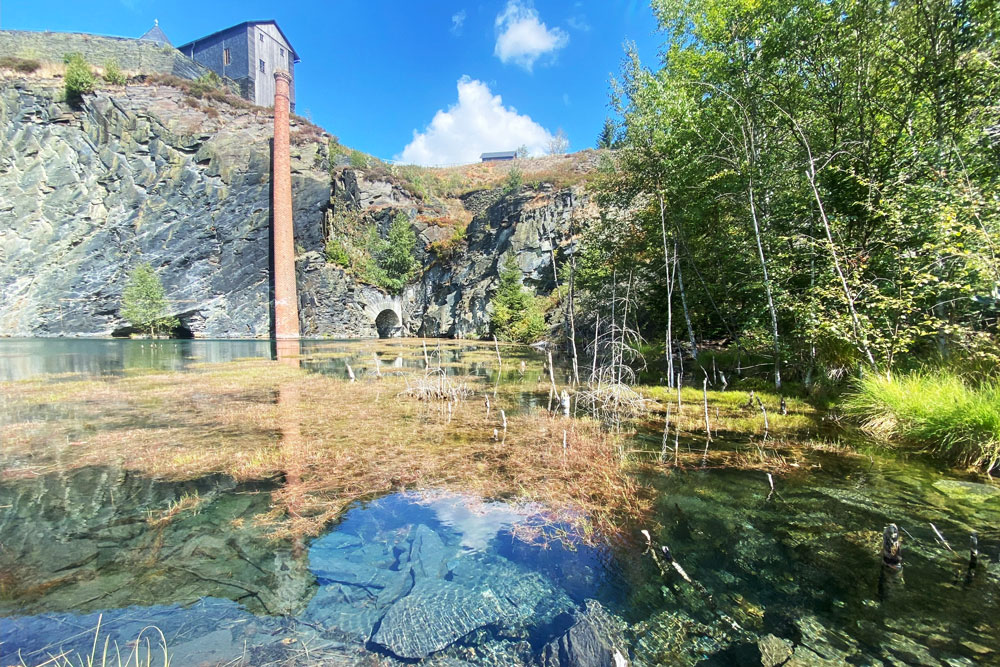 Klares Gewässer, in dem man Wasserpflanzen und Steine sehen kann, im Hintergrund eine Felswand mit einem Industrieturm, der aus der Wasser ragt.t