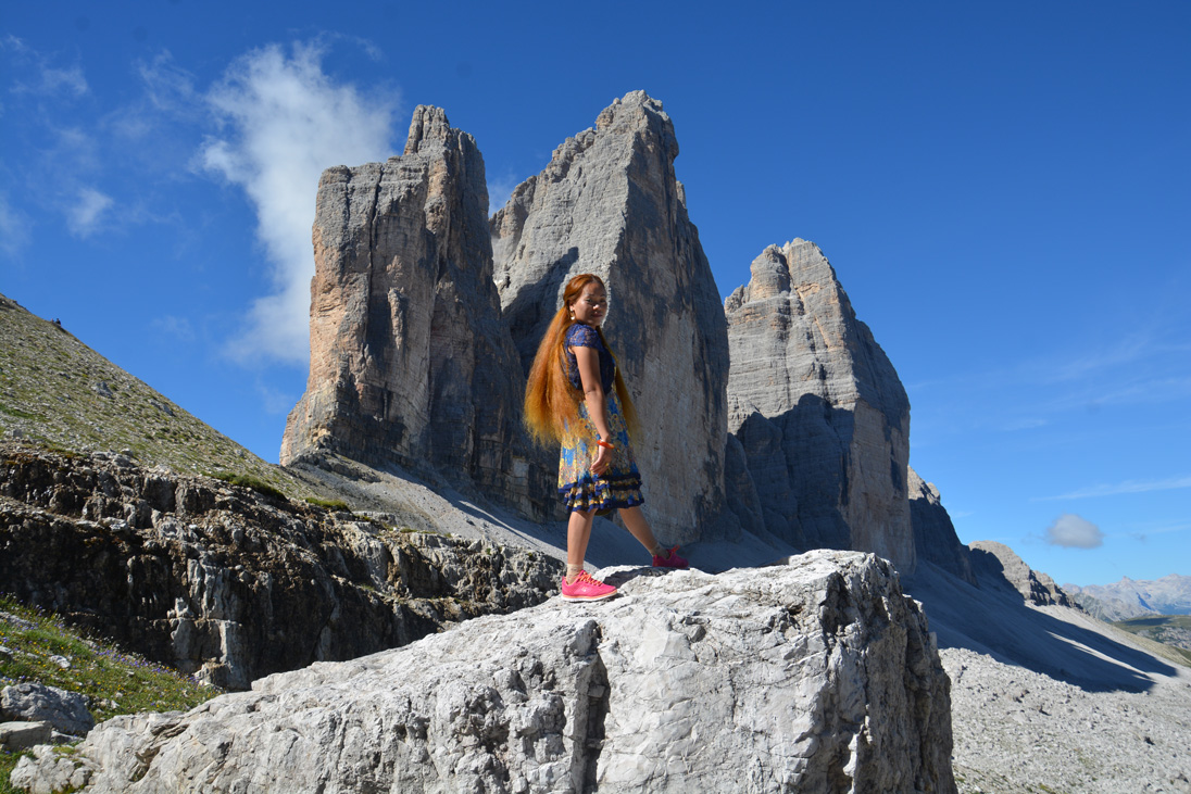 Eine Frau mit rotblonden Haaren posiert auf einem Fels vor einem imposanten Bergmassiv im Hintergrund