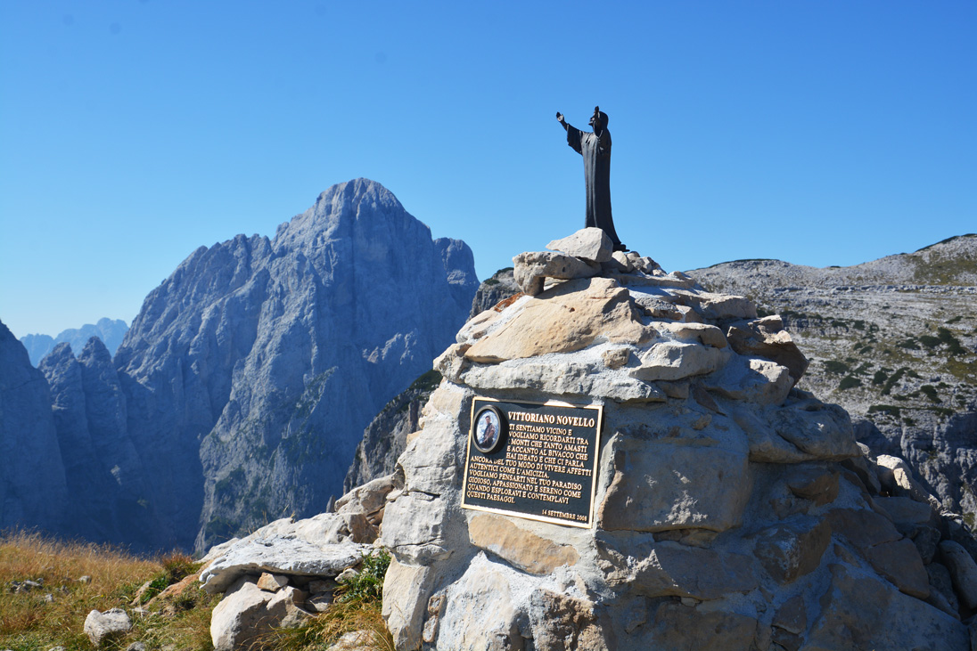 Eine Kunstfigur steht auf einem Steinhaufen mit einer Texttafel, im Hintergrund weitere Berge