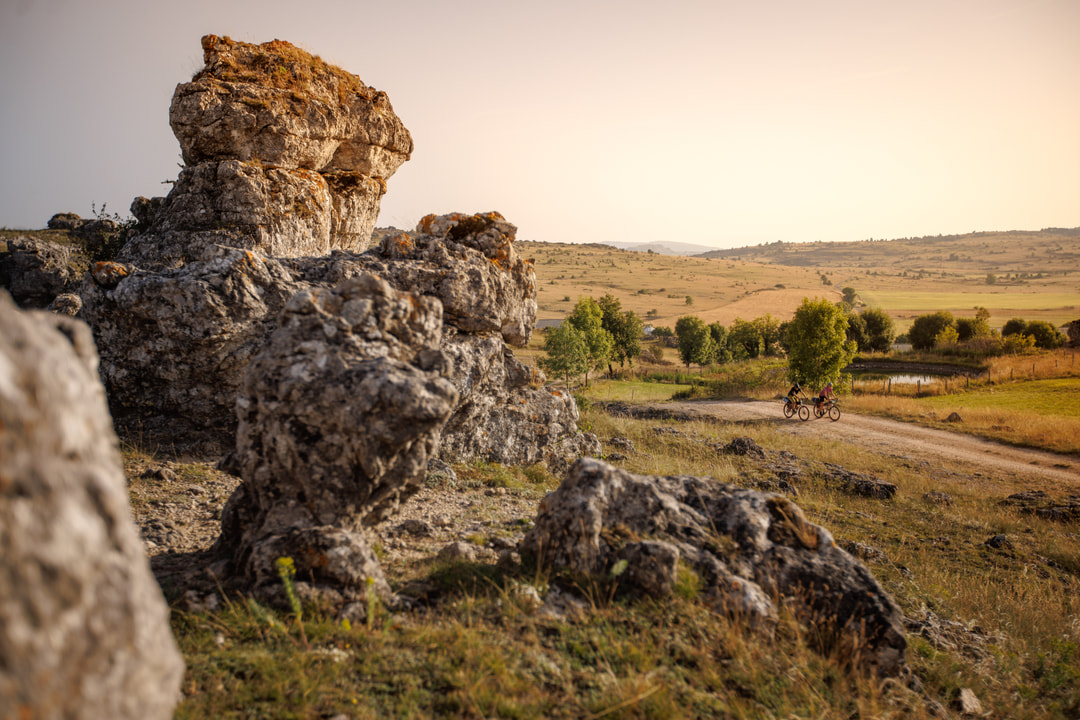 Zwei Radfahrende in einer goldgelben, hügeligen Hochebene mit markanten Felsen im Vordergrund