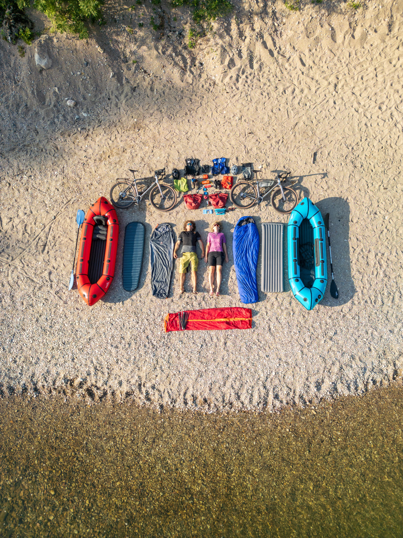 Luftaufnahme von zwei Personen inmitten ihrer ausgebreiteten Ausrüstungsgegenstände auf dem sandigen Flussufer