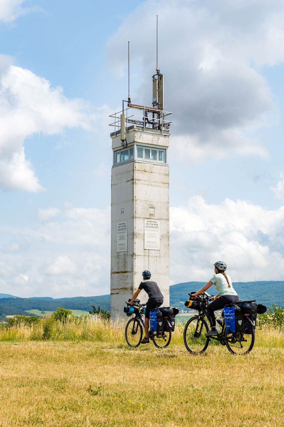 Zwei Radfahrende radeln an einem Grenzturm vorbei