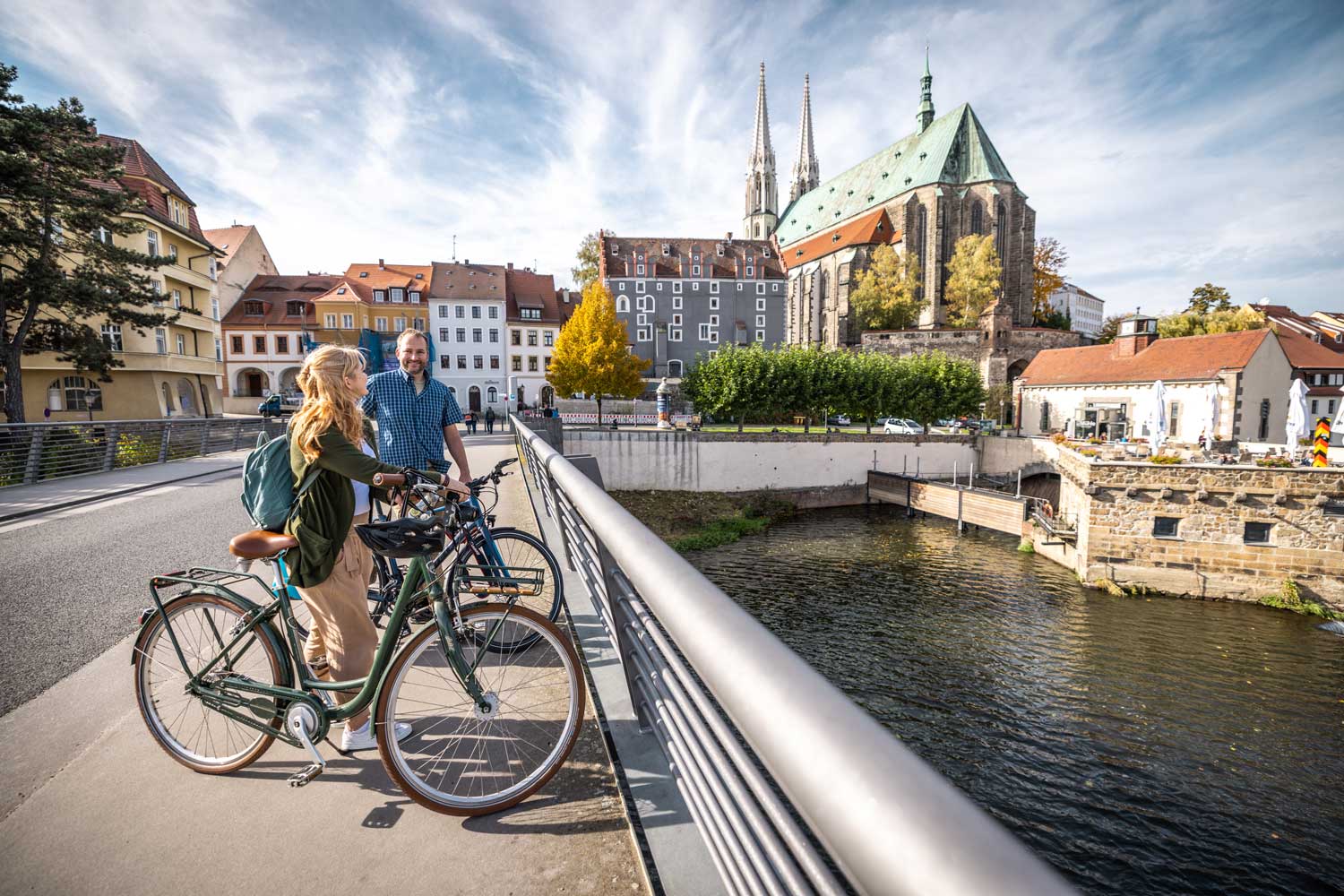 Zwei Radfahrende an einem Fluss, im Hintergrund eine Stadt