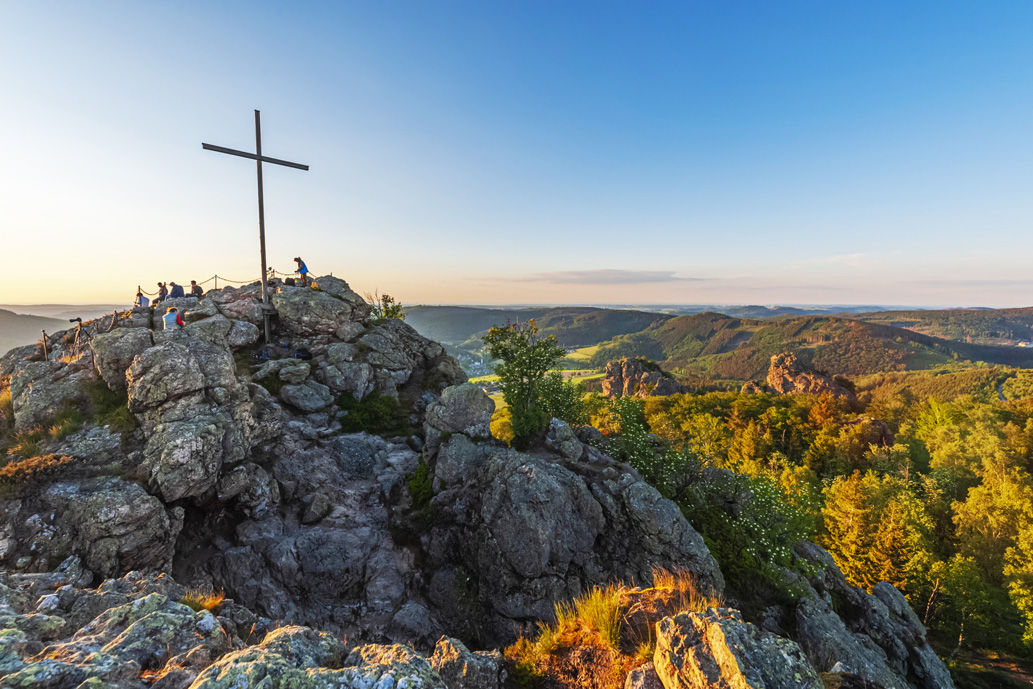 Ein Felsgipfel mit Gipfelkreuz inmitten einer hügeligen Waldlandschaft